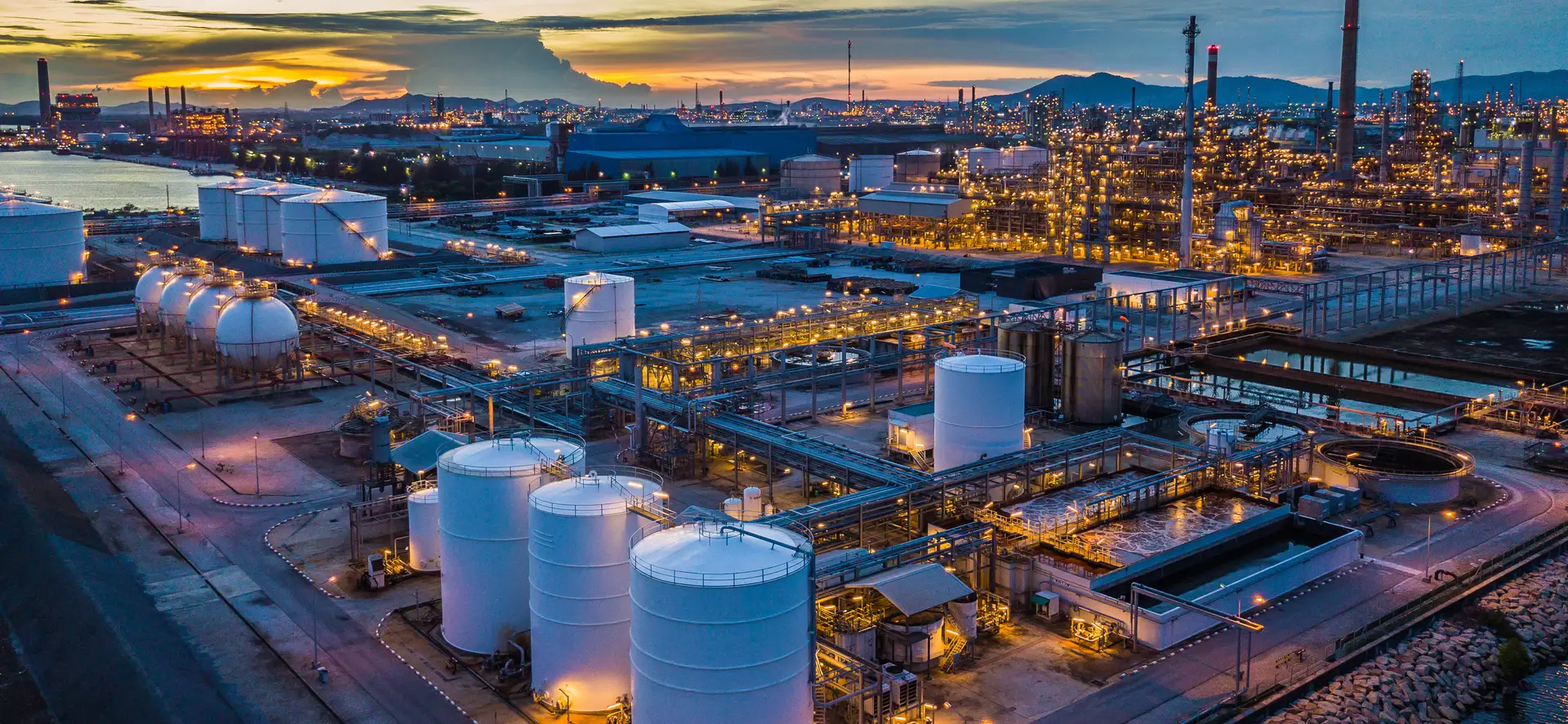 Aerial view of a lit industrial processing plant and tank farm at dusk, with a refinery skyline behind it