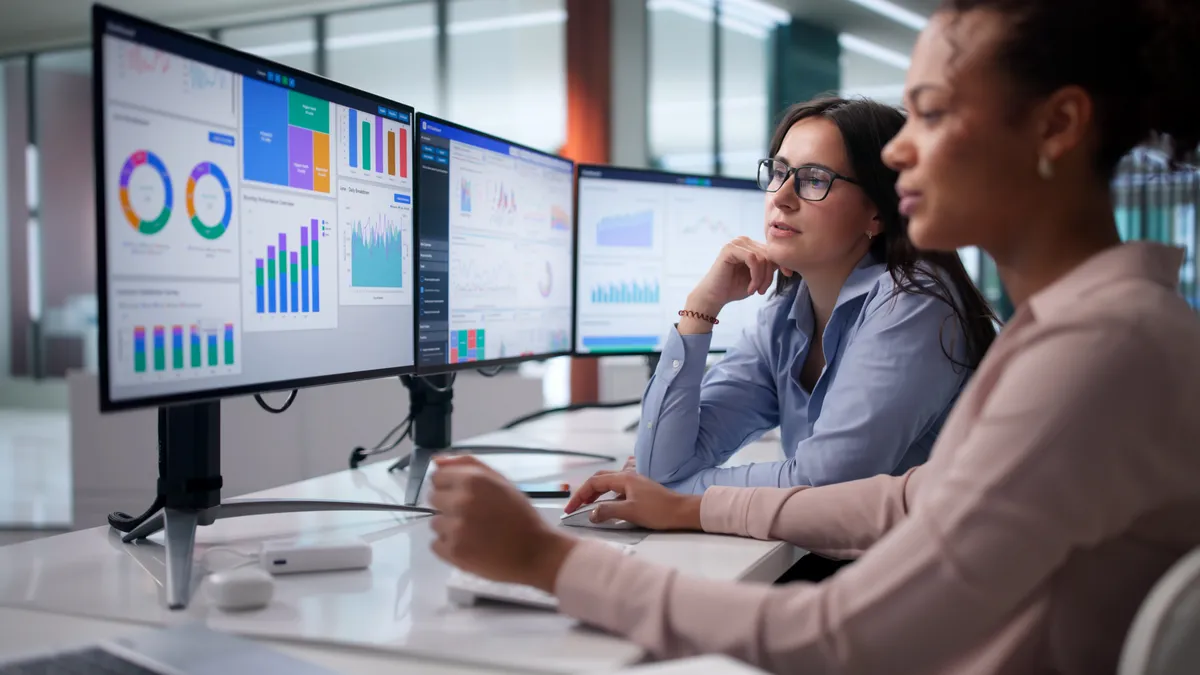 Two consultants at a desk reviewing workforce and labor-cost dashboards across three monitors.